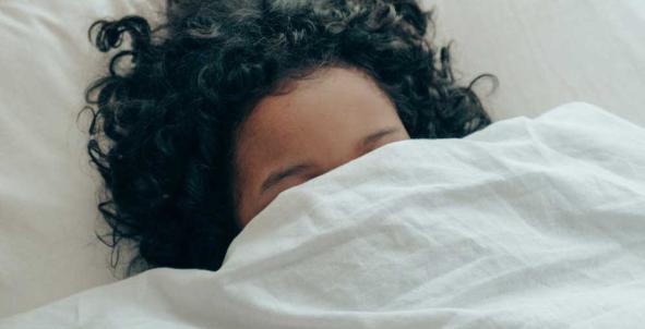 Een vrouw ligt onder de dekens in bed, alleen haar haar en voorhoofd is zichtbaar