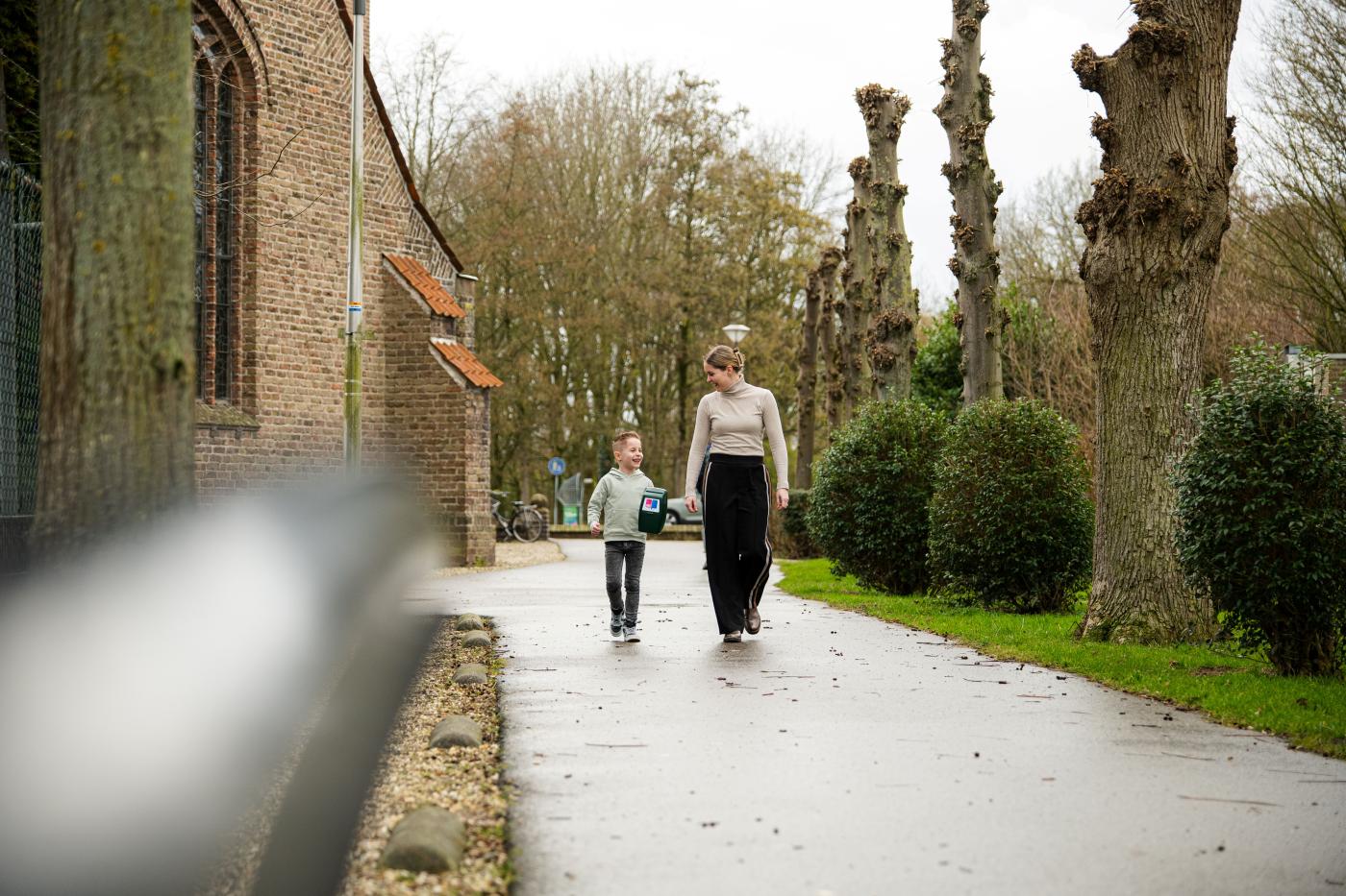 Moeder en kind lopen door een straat met een groene collectebus van Longfonds